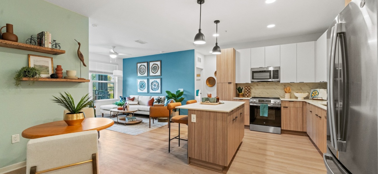 An interior view of a luxury apartment in Ocoee, FL at Elan Crown Point showcasing a vibrantly decorated living room, dining area, and kitchen. The living room features a bright blue back wall decorated with four paintings, has a couch, center table, side