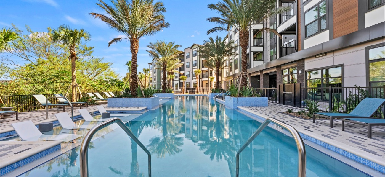 An exterior view of Elan Crown Point's courtyard at its luxury apartments in Ocoee, FL featuring a resort-inspired pool with pool railings and an array of poolside lounge chairs, palm trees, and plants, and a gated fence section for entering and exiting t